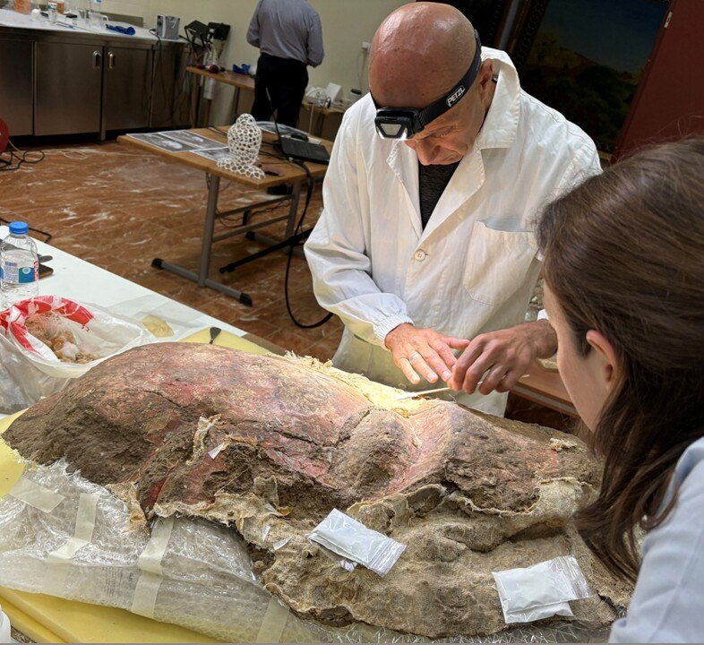 Two people lean over an artifact while one uses a tool to gently scrape the top layer.