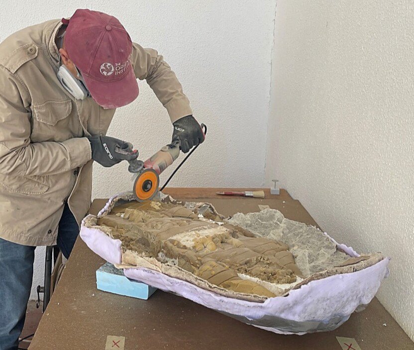 A person wearing protective gear uses a compact, circular saw to carve along the edge of a large fragment covered in a white substance.