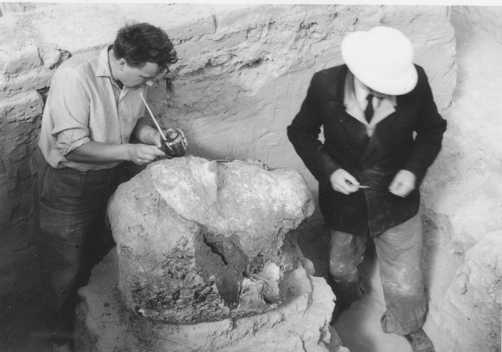 Two people with tools and protective equipment work on a large sculpture of a head in an excavation site.