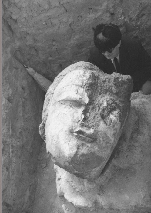 A large face emerges from stone in an excavation site as a man wearing a suit stands beside it.