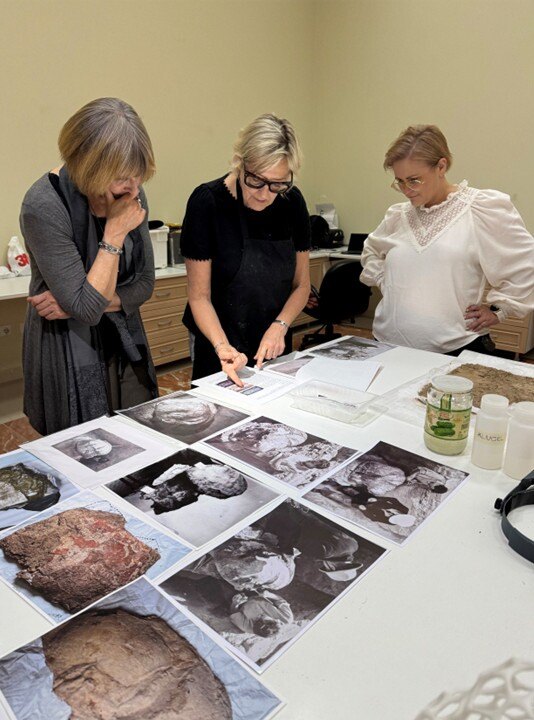 Three women analyze photographs laid out on a table.