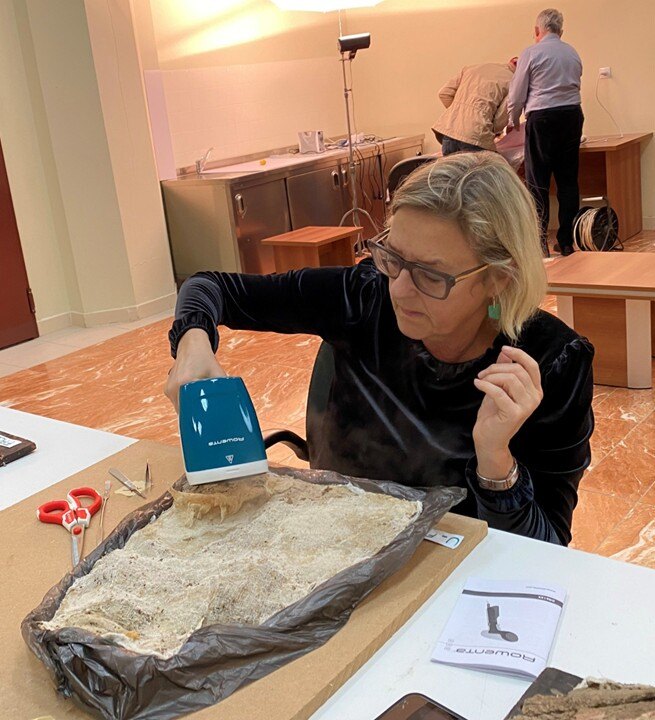 A woman holds a steamer over a fragment in a work room.