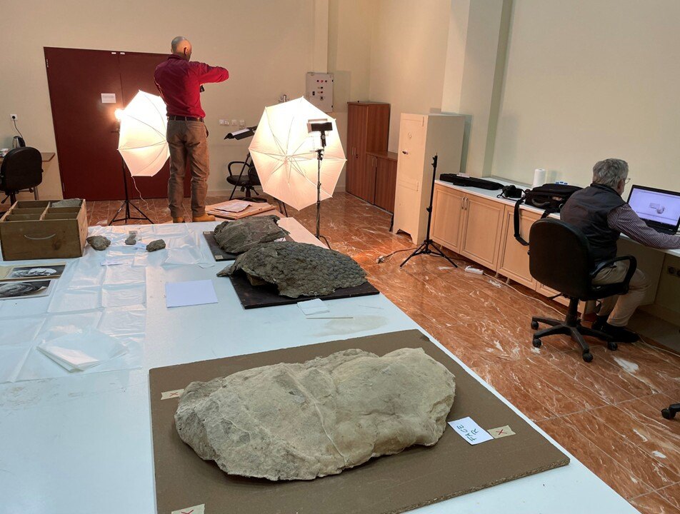Large, labeled fragments sit on a table in a work room with people working on computers and setting up lighting.