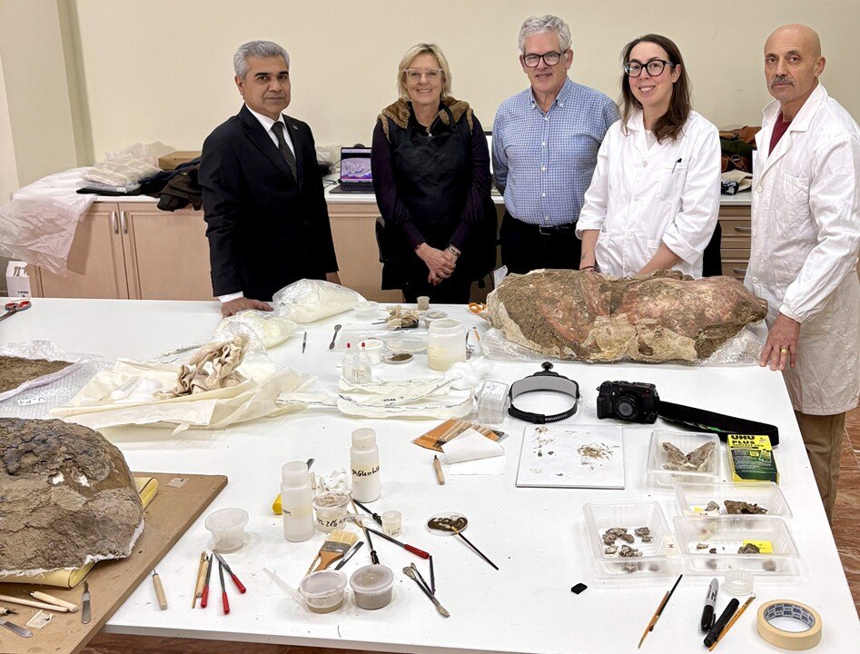 A group of five people smile for a photo in front of a large artifact surrounded by tools on a table.