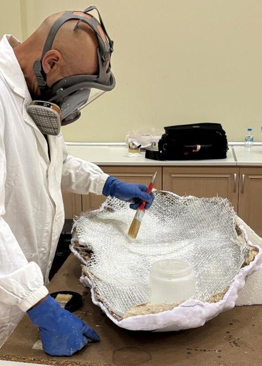 A worker wearing protective gear uses a paintbrush on a large artifact covered in gauze.